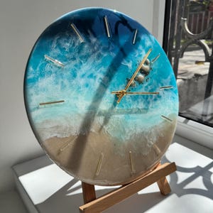 May include: A round clock with a beach-themed design. The clock face features a gradient of blue and white, resembling ocean waves, with a sandy-colored base. Gold-colored hands and markers indicate the time. The clock is displayed on a small wooden easel.