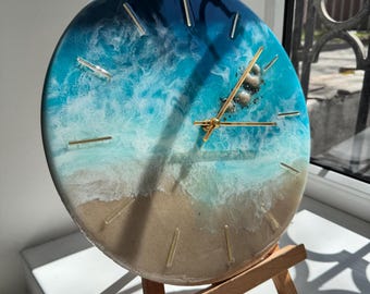 May include: A round clock with a beach-themed design. The clock face features a gradient of blue and white, resembling ocean waves, with a sandy-colored base. Gold-colored hands and markers indicate the time. The clock is displayed on a small wooden easel.