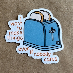May include: A sticker featuring a blue toaster with two slices of toast popping out. The sticker has the text "I want to make things even if nobody cares" in brown letters.