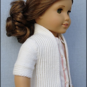 Könnte beinhalten: Ein weißer Strickcardigan mit gerippter Struktur und Knopfleiste. Der Cardigan wird von einer Puppe mit braunen Haaren getragen.