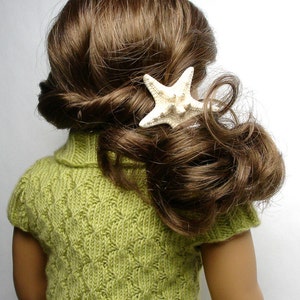 Pu&ograve; includere: Una bambola con lunghi capelli castani che indossa un maglione di lana verde con un fermaglio per capelli a forma di stella marina bianca.