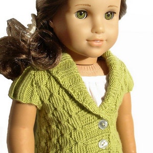 Pu&ograve; includere: Una bambola che indossa un cardigan di maglia verde con collo e bottoni. Il cardigan ha un motivo a treccia e viene indossato sopra una camicia bianca.