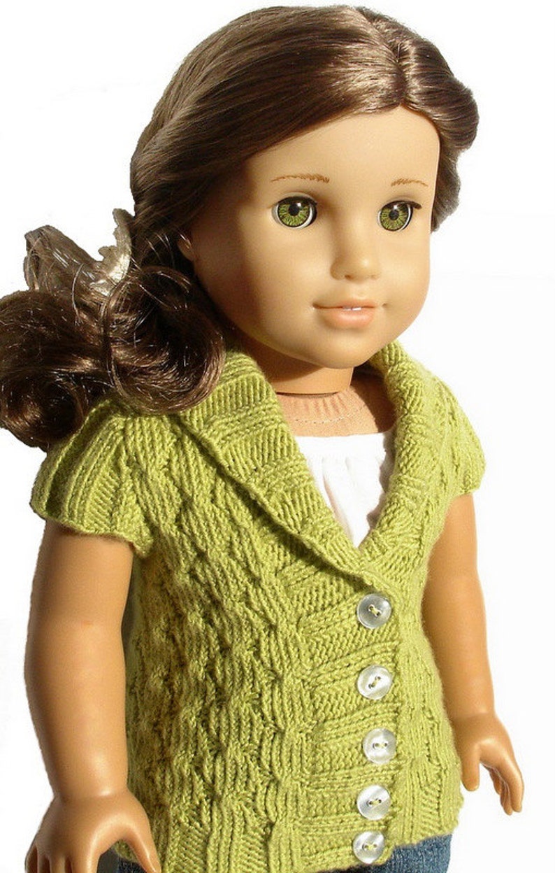 Pu&ograve; includere: Una bambola che indossa un cardigan di maglia verde con collo e bottoni. Il cardigan ha un motivo a treccia e viene indossato sopra una camicia bianca.
