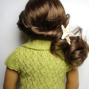 Pu&ograve; includere: Un maglione di lana verde con un motivo a treccia e maniche corte. Il maglione ha un collo alto ed &egrave; indossato da una bambola con i capelli castani.
