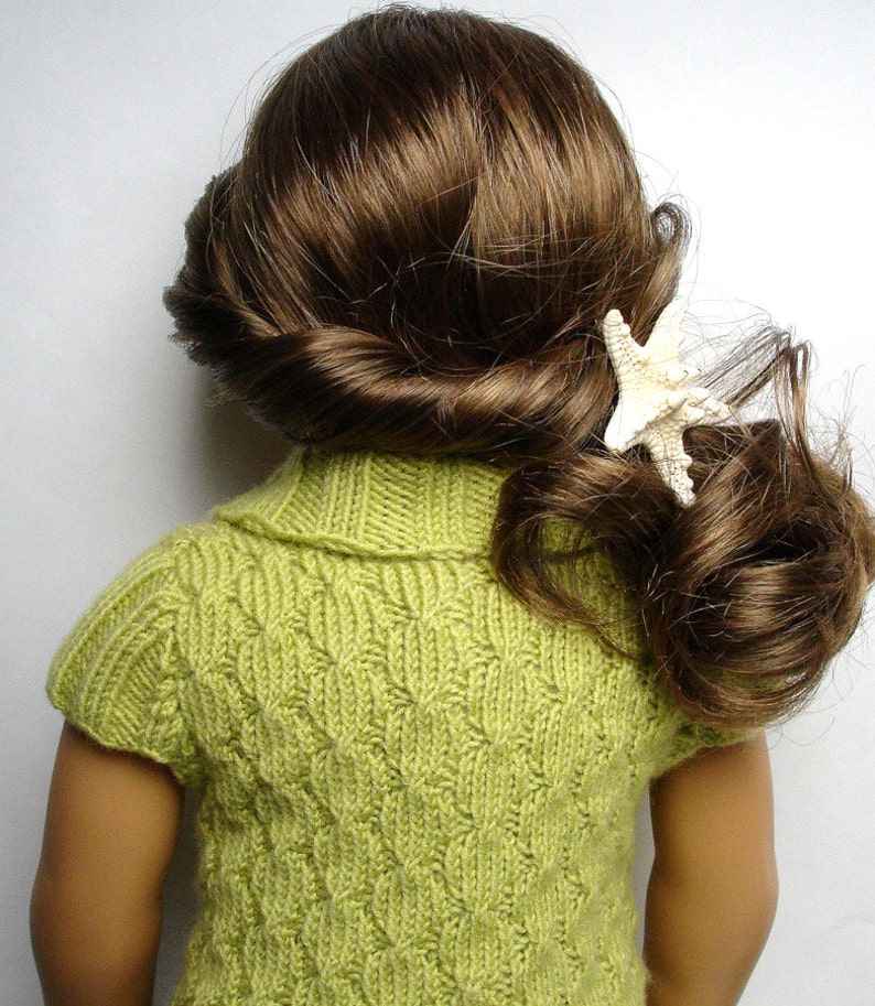 Pu&ograve; includere: Un maglione di lana verde con un motivo a treccia e maniche corte. Il maglione ha un collo alto ed &egrave; indossato da una bambola con i capelli castani.