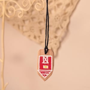 Puede incluir: Un colgante de madera con forma de escudo, suspendido de un cordón negro fino. La parte frontal presenta un rectángulo rojo pintado con un borde blanco, un pequeño cuadrado amarillo y una base triangular blanca con un patrón en zigzag.