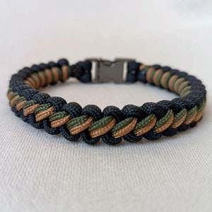 Könnte beinhalten: Ein geflochtenes Armband in Schwarz und Braun mit einem Metallverschluss. Das Armband ist aus Paracord und hat ein Tarnmuster.