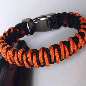 Puede incluir: Un brazalete de paracord negro y naranja con un cierre plateado.
