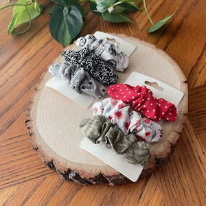 May include: A collection of fabric scrunchies in various patterns and colors, including black and white floral, gray with white dots, red with white polka dots, and green floral. The scrunchies are displayed on white cardboard tags, arranged on a wooden slice.