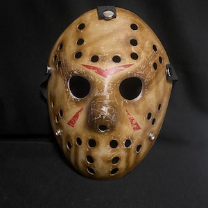 May include: A tan hockey-style mask with a weathered, distressed finish featuring numerous circular ventilation holes. The mask displays red triangular markings above and below the eye openings, with silver-toned metal rivets securing black straps.
