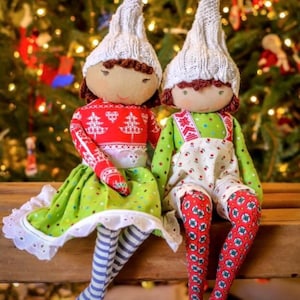Peut inclure: Deux poupées de Noël faites à la main sont assises côte à côte. L'une porte un pull rouge avec un motif d'arbre blanc, une jupe verte et des bas rayés. L'autre porte une tenue verte avec des bas à motifs rouges. Les deux poupées ont des chapeaux en tricot blanc.