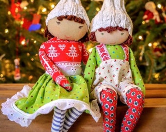 Pode incluir: Duas bonecas de Natal feitas à mão estão sentadas lado a lado. Uma boneca usa um suéter vermelho com um desenho de árvore branca, uma saia verde e meias listradas. A outra boneca usa uma roupa verde com meias estampadas vermelhas. Ambas as bonecas têm chapéus de malha brancos.