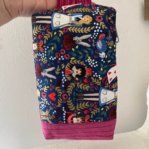 May include: A small blue fabric bag with a pink handle. The bag is decorated with a whimsical pattern of Alice in Wonderland characters and floral designs.