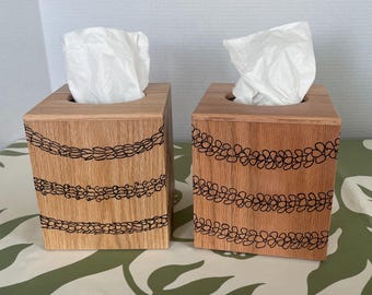 May include: Two wooden tissue box covers with a floral design. The tissue boxes are made of light brown wood and have a black floral design.