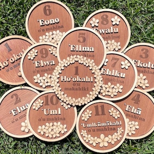 May include: A collection of eleven wooden coasters with numbers and Hawaiian words. Each coaster is round with a light wood border and a darker wood center. The numbers are accompanied by floral embellishments and text.