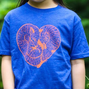 May include: A blue t-shirt featuring an orange heart-shaped design with two foxes. The foxes are intertwined, creating the heart shape. The shirt has short sleeves and a crew neck.