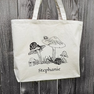 Op de afbeelding: Een witte canvas tote bag met zwarte lijntekeningen van paddenstoelen, slakken en een vlinder. De tas heeft de naam "Stephanie" op de voorkant gedrukt.