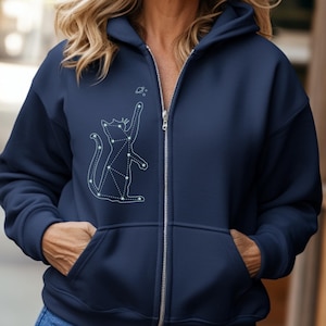 Puede incluir: Sudadera con capucha azul marino con cremallera y diseño de constelación de gato. El gráfico blanco representa un gato que alcanza un pequeño planeta. La sudadera tiene cremallera completa, capucha y dos bolsillos delanteros.