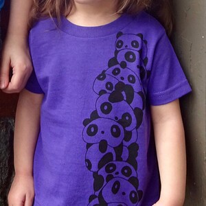K&ouml;nnte beinhalten: Ein junges M&auml;dchen tr&auml;gt ein lila T-Shirt mit einem schwarzen und wei&szlig;en Panda-Stapel-Motiv. Das Shirt hat kurze &Auml;rmel und einen Rundhalsausschnitt.
