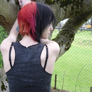 Pode incluir: Uma pessoa com cabelo preto e rosa est&aacute; vestindo um top cinza sem mangas com al&ccedil;as pretas. Ela est&aacute; em p&eacute; na frente de uma cerca de arame e uma &aacute;rvore.