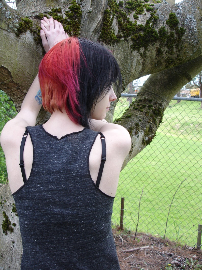 Pode incluir: Uma pessoa com cabelo preto e rosa est&aacute; vestindo um top cinza sem mangas com al&ccedil;as pretas. Ela est&aacute; em p&eacute; na frente de uma cerca de arame e uma &aacute;rvore.