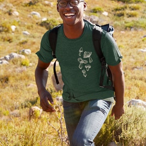 May include: A man wearing a green t-shirt with a white mushroom graphic. He is smiling and walking on a grassy hill.