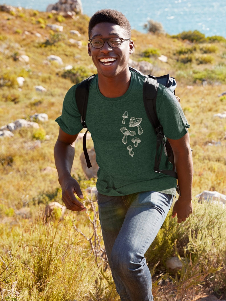 May include: A man wearing a green t-shirt with a white mushroom graphic. He is smiling and walking on a grassy hill.