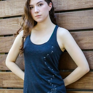 Pode incluir: Uma mulher vestindo uma camiseta sem mangas azul escura com um desenho de dente-de-le&atilde;o branco. A camiseta sem mangas tem um decote redondo e um caimento solto.