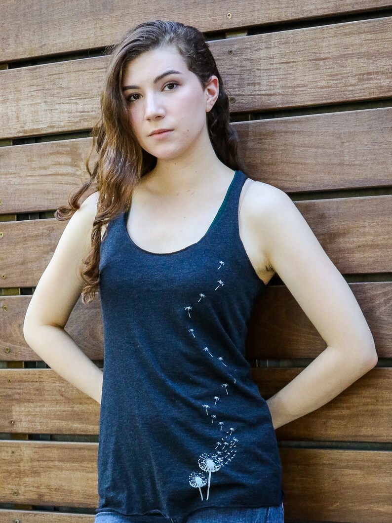 Pode incluir: Uma mulher vestindo uma camiseta sem mangas azul escura com um desenho de dente-de-le&atilde;o branco. A camiseta sem mangas tem um decote redondo e um caimento solto.