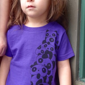 K&ouml;nnte beinhalten: Ein junges M&auml;dchen tr&auml;gt ein lila T-Shirt mit einem schwarz-wei&szlig;en Panda-Druck. Das Shirt zeigt einen Stapel Pandas mit sichtbaren Gesichtern. Das M&auml;dchen tr&auml;gt dunkelblaue Jeans mit wei&szlig;en Punkten.