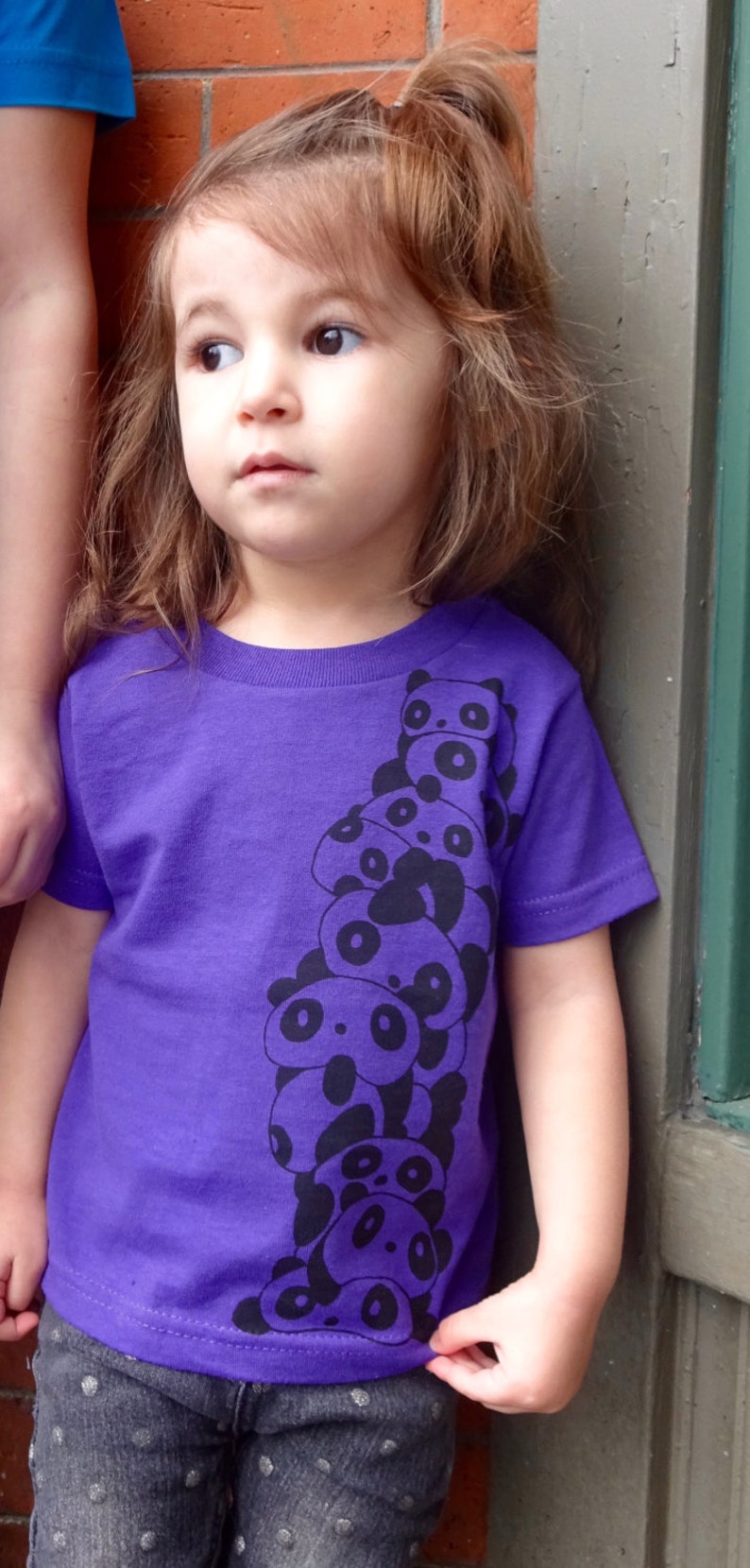 K&ouml;nnte beinhalten: Ein junges M&auml;dchen tr&auml;gt ein lila T-Shirt mit einem schwarz-wei&szlig;en Panda-Druck. Das Shirt zeigt einen Stapel Pandas mit sichtbaren Gesichtern. Das M&auml;dchen tr&auml;gt dunkelblaue Jeans mit wei&szlig;en Punkten.