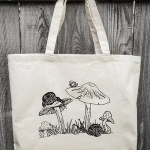 Op de afbeelding: Een witte canvas tote bag met een zwart-witte illustratie van paddenstoelen en slakken. De illustratie toont drie paddenstoelen, twee met slakken erop, en een vlinder.