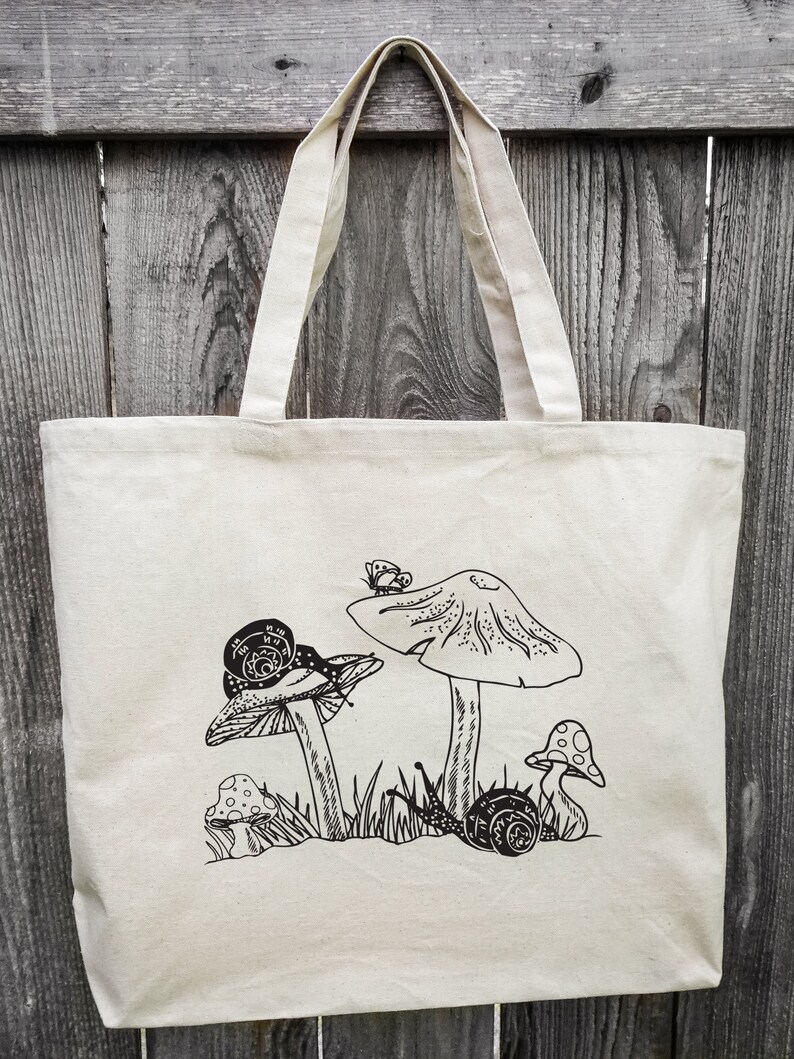 Op de afbeelding: Een witte canvas tote bag met een zwart-witte illustratie van paddenstoelen en slakken. De illustratie toont drie paddenstoelen, twee met slakken erop, en een vlinder.