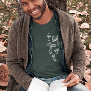 May include: A man wearing a green t-shirt with a white mushroom graphic. The t-shirt has a crew neck and short sleeves. The man is sitting on the ground with a book in his lap. He is wearing a grey beanie and a brown cardigan.