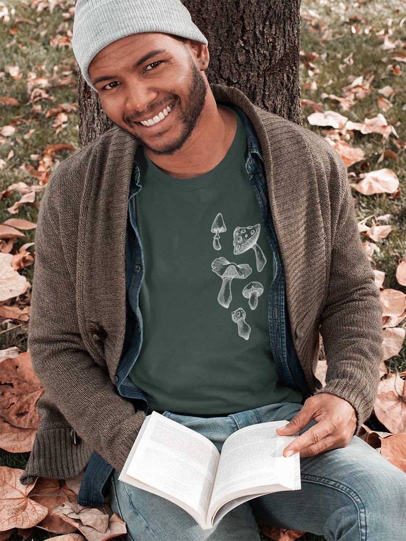 May include: A man wearing a green t-shirt with a white mushroom graphic. The t-shirt has a crew neck and short sleeves. The man is sitting on the ground with a book in his lap. He is wearing a grey beanie and a brown cardigan.