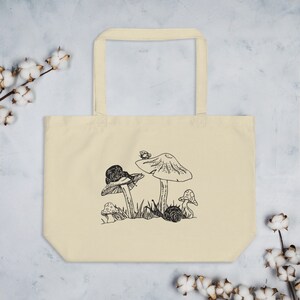 Op de afbeelding: Een beige tote bag met een zwart-witte illustratie van paddenstoelen en slakken. De illustratie toont drie paddenstoelen met een slak op elke paddenstoel. Er zit een kleine vlinder op de top van een van de paddenstoelen.