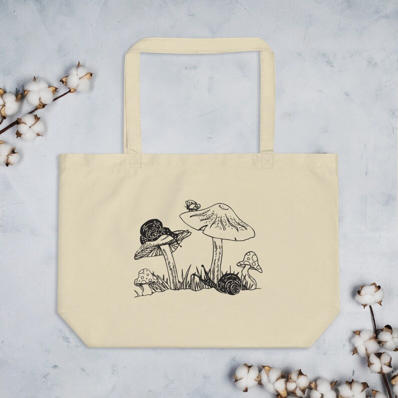 Op de afbeelding: Een beige tote bag met een zwart-witte illustratie van paddenstoelen en slakken. De illustratie toont drie paddenstoelen met een slak op elke paddenstoel. Er zit een kleine vlinder op de top van een van de paddenstoelen.
