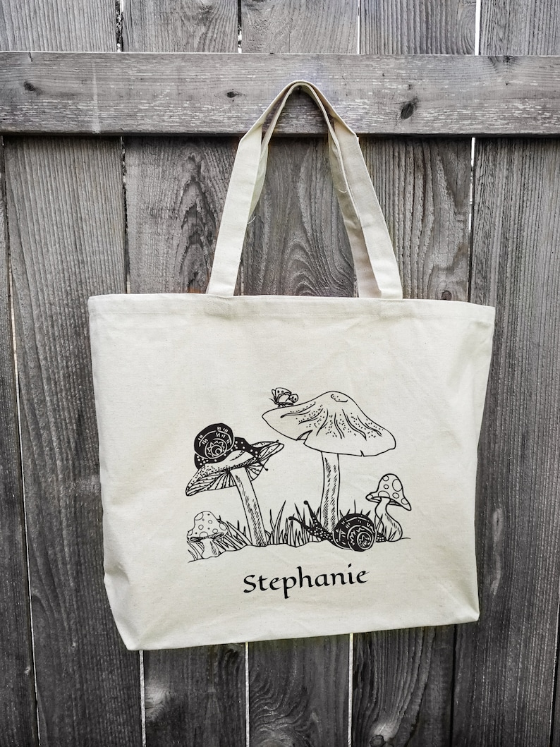 Op de afbeelding: Een witte canvas tote bag met zwarte lijntekeningen van paddenstoelen, slakken en een vlinder. De tas heeft de naam "Stephanie" op de voorkant gedrukt.