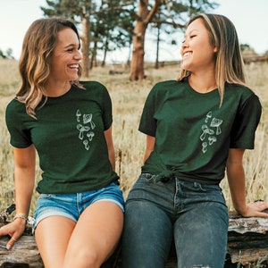 May include: Two women wearing dark green t-shirts with a white mushroom graphic. The shirts have short sleeves and a crew neck. The women are sitting on a log in a forest setting.