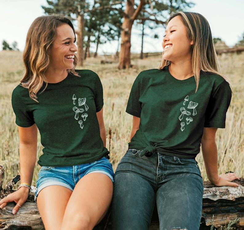 May include: Two women wearing dark green t-shirts with a white mushroom graphic. The shirts have short sleeves and a crew neck. The women are sitting on a log in a forest setting.