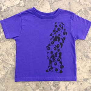 K&ouml;nnte beinhalten: Lilafarbenes T-Shirt mit kurzen &Auml;rmeln und einem schwarzen Aufdruck einer Stapel von Pandas.