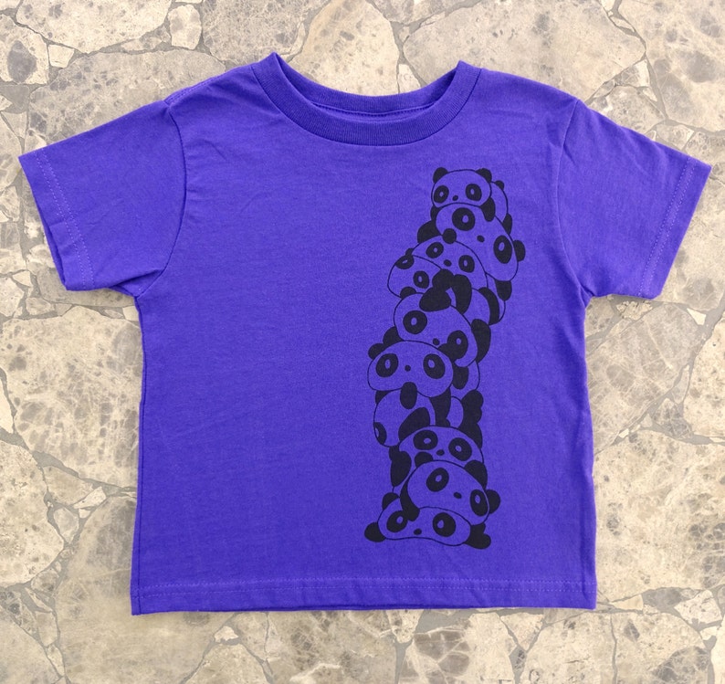 K&ouml;nnte beinhalten: Lilafarbenes T-Shirt mit kurzen &Auml;rmeln und einem schwarzen Aufdruck einer Stapel von Pandas.