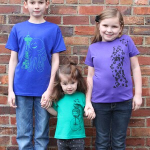 K&ouml;nnte beinhalten: Drei Kinder tragen bunte T-Shirts mit grafischen Designs. Der Junge links tr&auml;gt ein blaues Shirt mit einem gr&uuml;nen Oktopus und einem Turmmuster. Das M&auml;dchen in der Mitte tr&auml;gt ein gr&uuml;nes Shirt mit einer schwarz-wei&szlig;en Grafik einer Person, die ein Einrad f&auml;hrt. Das M&auml;dchen rechts tr&auml;gt ein lilafarbenes Shirt mit einer schwarz-wei&szlig;en Grafik eines Stapels Pandas.