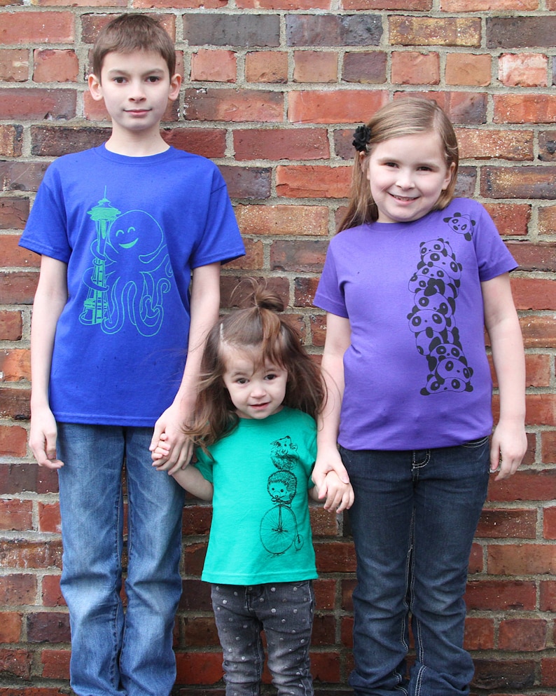 K&ouml;nnte beinhalten: Drei Kinder tragen bunte T-Shirts mit grafischen Designs. Der Junge links tr&auml;gt ein blaues Shirt mit einem gr&uuml;nen Oktopus und einem Turmmuster. Das M&auml;dchen in der Mitte tr&auml;gt ein gr&uuml;nes Shirt mit einer schwarz-wei&szlig;en Grafik einer Person, die ein Einrad f&auml;hrt. Das M&auml;dchen rechts tr&auml;gt ein lilafarbenes Shirt mit einer schwarz-wei&szlig;en Grafik eines Stapels Pandas.