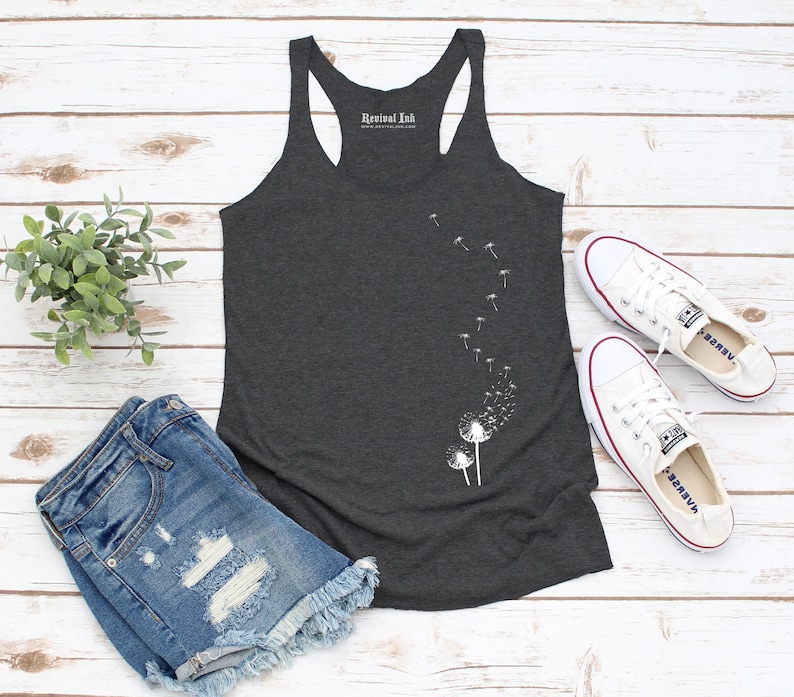 Pode incluir: Um top de al&ccedil;as cinza escuro com um desenho de dente-de-le&atilde;o branco e o texto "Revival Ink" nas costas. O top est&aacute; sobre uma superf&iacute;cie de madeira branca com um par de sapatos de lona brancos e um par de shorts jeans azuis.