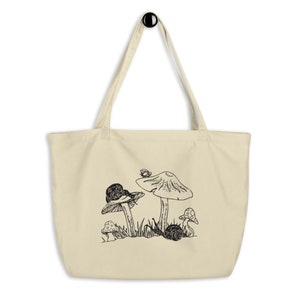 Op de afbeelding: Een beige tote bag met een zwart-witte illustratie van paddenstoelen en slakken. De illustratie toont een grote paddenstoel met een slak die erop kruipt, een kleinere paddenstoel met een slak erop en een vlinder die boven de grote paddenstoel vliegt.