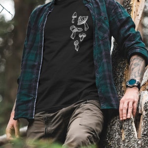 May include: A black t-shirt with a white graphic of mushrooms on the front. The shirt is being worn by a person standing in a forest.