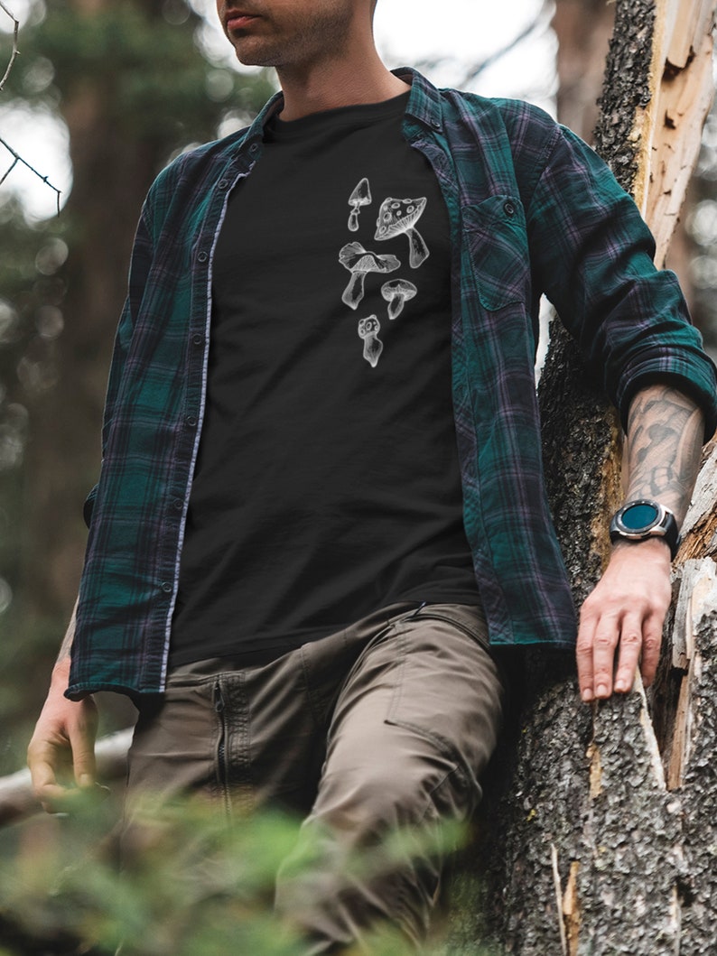 May include: A black t-shirt with a white graphic of mushrooms on the front. The shirt is being worn by a person standing in a forest.