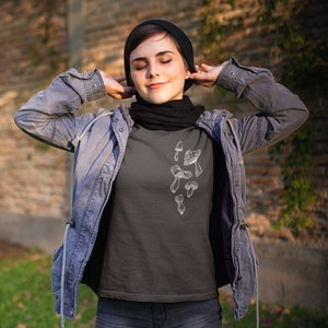 May include: A dark gray t-shirt with a white graphic of mushrooms. The person wearing the shirt is wearing a black beanie and a blue denim jacket.