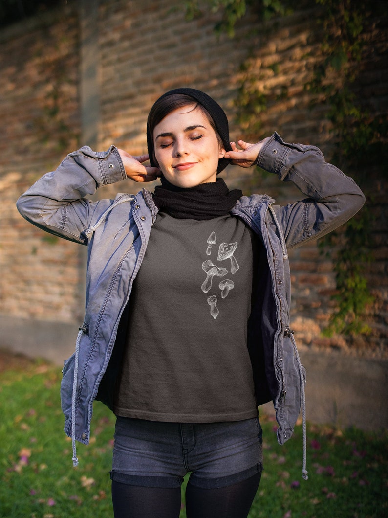 May include: A dark gray t-shirt with a white graphic of mushrooms. The person wearing the shirt is wearing a black beanie and a blue denim jacket.